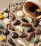 Assortiment de friture de Pâques en chocolat noir, lait et blanc disposé sur une table en bois avec un petit panier artisanal et des fleurs des champs