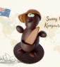 Montage de Pâques artisanal en chocolat représentant Sunny le Kangourou avec une casquette et une planche de surf, sur un socle en chocolat noir.